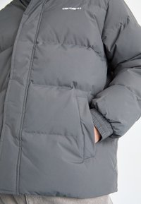Chaqueta acolchada gris con un acabado texturizado, que presenta un cierre de cremallera en la parte frontal, puños elásticos y un logo en el pecho. Dos bolsillos laterales visibles.