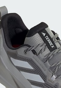 Zapatilla deportiva gris con una parte superior texturizada, que presenta acentos reflectantes, un collar acolchado y una etiqueta con el logotipo. Incluye cordones grises y una superficie con patrón.
