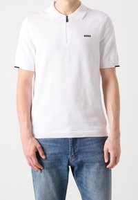 Polo blanc à manches courtes avec col zippé, logo noir sur la poitrine, et bordure noire contrastante sur les manches. Associé à un jean bleu.