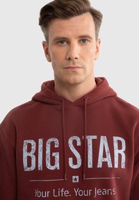 Bordeauxfarbener Hoodie mit Kordelzugkapuze. Die Vorderseite zeigt großen, abgenutzten weißen Text: "BIG STAR" und kleineren Text "Dein Leben. Deine Jeans."