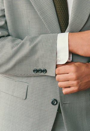 Mann richtet den Ärmelaufschlag eines Sakkos mit Hahnentrittmuster, sichtbar sind weißes Hemd und dunkle, strukturierte Krawatte.