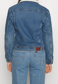 Džínová bunda v středně modré barvě s beránkovým límcem, přiléhavý střih a viditelné švy. Hnědá kožená nášivka na zadní straně blízko pasu.