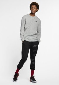 Camisa cinza de mangas compridas com o logótipo da Nike em preto, combinada com calças de moletom pretas com logótipos da Nike repetidos em preto. Sapatilhas pretas e vermelhas.