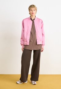 Giacca bomber in satin rosa sopra una tunica marrone, abbinata a pantaloni larghi marroni e sneakers bianche con accenti rossi su un pavimento giallo.