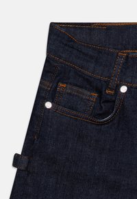 Tmavě modré džíny z denimu, detailní záběr na přední kapsu, malou kapsičku na mince, oranžové šití a kovové nýty na bílém pozadí.