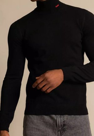 Homme portant un pull noir à col roulé ajusté avec un petit logo rouge "HUGO" au niveau du cou, associé à un pantalon gris.