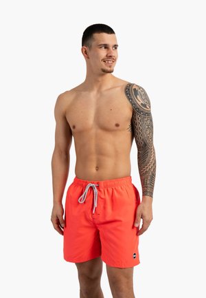 Homme musclé torse nu avec un tatouage tribal à manches longues, portant un short de bain orange vif, regardant de côté avec un léger sourire sur fond blanc.