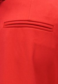 Κοντινή λήψη κόκκινου τσέπης ρούχου με οριζόντια σχισμή και ραμμένες λεπτομέρειες σε λείο ύφασμα.