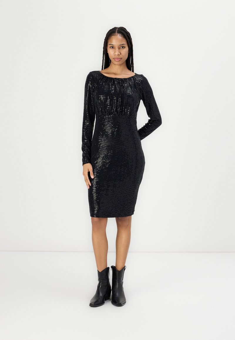 Czarna sukienka z cekinami, z długimi rękawami, dopasowaną sylwetką i marszczonym dekoltem. Noszona z czarnymi botkami na kostkę na neutralnym tle.