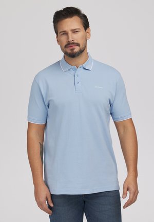 Mężczyzna z krótkimi ciemnymi włosami i brodą, ubrany w jasnoniebieską koszulkę polo i dżinsy, stojący na jednolitym szarym tle.