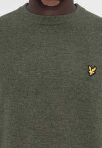 Pull en vert olive avec un col rond, texture tricotée, et un petit logo d'oiseau noir et jaune sur la poitrine.