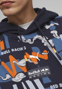 Felpa con cappuccio navy che presenta motivi astratti bianchi e arancioni, un logo Puma e un'etichetta Red Bull Racing sul davanti.