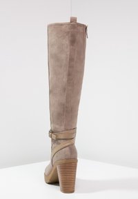Bota de ante en taupe hasta la rodilla con un tacón ancho y apilado, cremallera lateral y un detalle de correa decorativa alrededor del tobillo. Suela de goma texturizada.