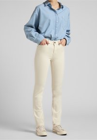 Persona con camisa de manga larga azul claro, jeans ajustados color blanco roto y zapatillas color beige, de pie con las manos en los bolsillos sobre un fondo gris.