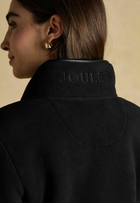 Žena s hnedými stredne dlhými vlasmi, ktorá má na sebe zlaté kruhové náušnice a čiernu fleecovú bundu s vyšitým nápisom "JOULES" na zadnom golieriku.