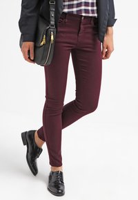 Person som bär burgundiska smala byxor, svarta oxfordskor, en rutmönstrad skjorta och en svart jacka, och håller en svart läderhandväska.