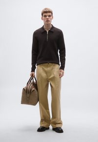 Καφέ πουλόβερ με φερμουάρ στον λαιμό, μπεζ παντελόνι φαρδύ στο πόδι, μαύρα παπούτσια, κρατώντας μια καφέ τσάντα duffel με καφέ λεπτομέρειες. Απλός και μοντέρνος σχεδιασμός.