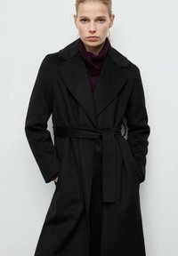 Czarny trencz z wełny z paskiem w talii, klapami z wycięciem i długimi rękawami. Noszony na dopasowanym fioletowym golfie, z bocznymi kieszeniami.