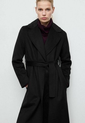 Czarny trencz z wełny z paskiem w talii, klapami z wycięciem i długimi rękawami. Noszony na dopasowanym fioletowym golfie, z bocznymi kieszeniami.