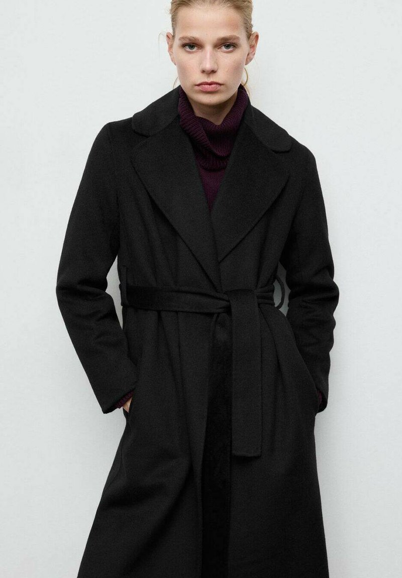 Czarny trencz z wełny z paskiem w talii, klapami z wycięciem i długimi rękawami. Noszony na dopasowanym fioletowym golfie, z bocznymi kieszeniami.