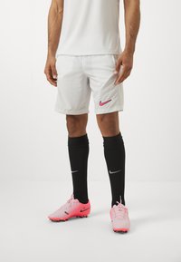 Fehér sportnadrág rózsaszín Nike logóval, fekete, térdig érő zoknival, amelyen ezüst Nike pipa található és rózsaszín focicipőkkel párosítva.