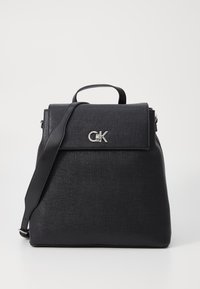 Sac à dos noir texturé de forme rectangulaire, avec une poignée supérieure, une bandoulière ajustable et un logo métallique à l'avant.