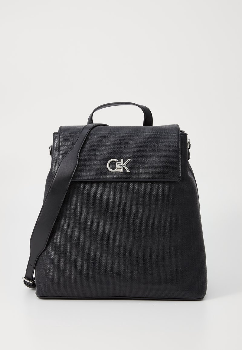 Sac à dos noir texturé de forme rectangulaire, avec une poignée supérieure, une bandoulière ajustable et un logo métallique à l'avant.