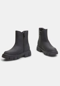Botas negras hasta el tobillo hechas de material suave, con cremallera lateral, suela de goma gruesa y diseño de puntera redonda.