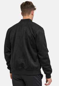 Zwarte faux-suède bomberjack met ribgebreide mouwen en onderkant, met een gladde textuur en een gestroomlijnd design, gezien vanaf de achterkant.