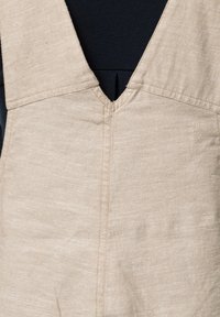 Beige linen jumpsuit with a deep V-neckline and wide straps. The fabric features a subtle texture with visible stitching details.