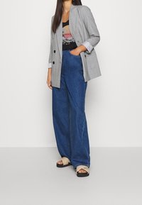 Blazer oversize à carreaux gris avec de larges revers, porté sur un débardeur noir à motif graphique, associé à un jean large en denim bleu taille haute et des sandales beiges.