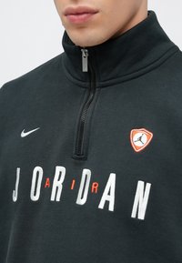 Μαύρο φούτερ με φερμουάρ και ψηλό γιακά, με λευκή επιγραφή "JORDAN" και κόκκι�νο τρίγωνο λογότυπο στο αριστερό στήθος. Μαλακή υφή.