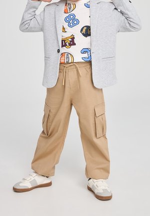 Barn iført beige cargobukser, hvide sneakers, en grå blazer og en hvid skjorte med blå og orange sportstema-prints.