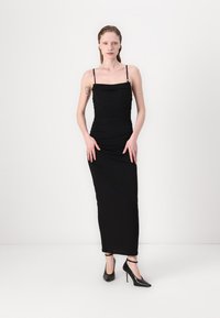 Černé, přiléhavé šaty až na zem s nařasenými detaily a tenkými ramínky. Modelka má na sobě černé vysoké podpatky. Hladký, elastický materiál.