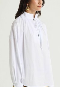 Blusa bianca con colletto alto, dettagli con coulisse e maniche lunghe gonfie. Realizzata in tessuto di cotone liscio per il massimo comfort.