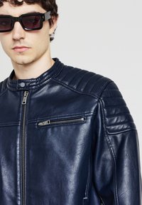 Veste en cuir bleu marine avec des accents côtelés aux épaules et une poche zippée. Comprend un col montant et une texture lisse. Modèle portant des lunettes de soleil sombres.