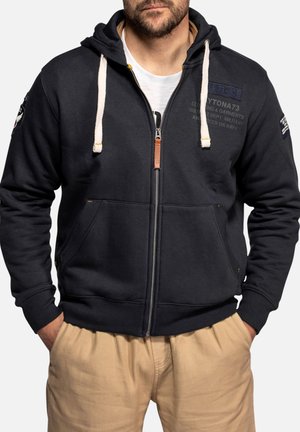 Mand iført sort lynlåshoodie med hvide snore og beige bukser, hænderne i lommerne, foran ensfarvet baggrund.