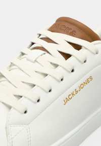Baskets blanches avec des lacets crème, une languette en cuir marron et un logo doré "JACK & JONES" sur le côté.