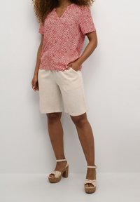 Žena má na sobě červený květinový top s krátkým rukávem, béžové šortky ke kolenům a béžové platformové sandály se zapínáním kolem kotníků, stojí před jednobarevným pozadím.