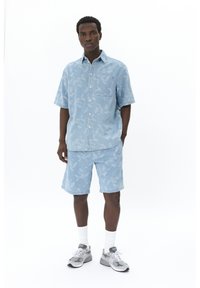 Camicia button-up in denim azzurro chiaro e pantaloni corti abbinati, con un motivo floreale, abbinati a sneakers grigie e calze bianche.