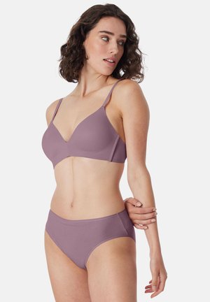 Femme portant un soutien-gorge et une culotte assortis mauve, regardant sur le côté, un bras plié avec la main reposant sur le coude opposé.