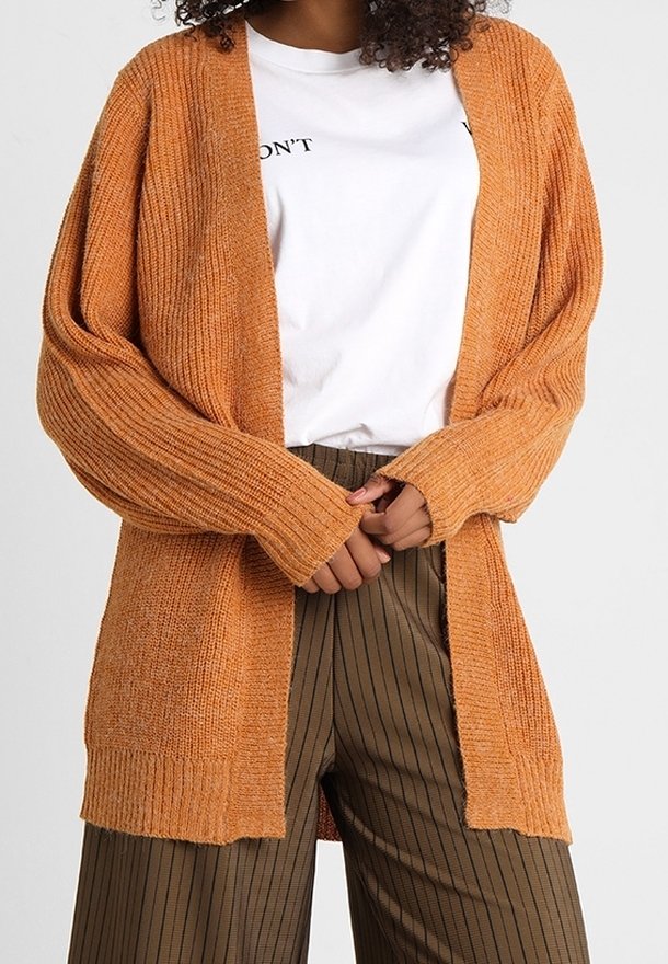 Cardigan de punto en color óxido con frente abierto, textura acanalada y mangas largas, llevado sobre una camiseta blanca y pantalones a rayas marrones.