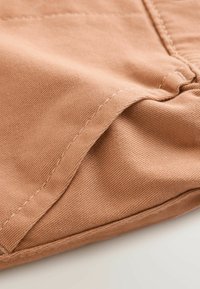 Light brown fabric swatch showing textured surface with visible stitching along the edges and seams. Soft cotton blend material.