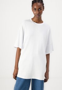 Bijela oversized majica kratkih rukava, okruglog izreza, izrađena od mekog pamučnog materijala, s opuštenim krojem i bočnim prorezima, nosi se uz plave traperice.