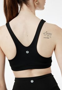 Czarny sportowy stanik z plecami w kształcie litery V, z logo na środku. Gładka tekstura i minimalistyczny design, w połączeniu z dopasowanymi legginsami z wysokim stanem.
