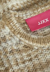 Pull en tricot beige et crème avec un motif texturé. Présente une étiquette rose avec "JJXX" en blanc. Gros plan sur les détails du tissu.