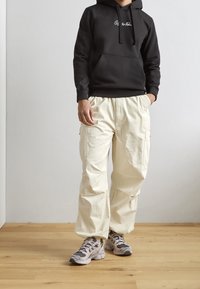 Felpa nera con cappuccio e testo bianco, abbinata a pantaloni cargo beige larghi e scarpe da ginnastica grigie. Sfondo in legno.