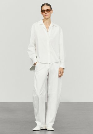 Woman with slicked-back hair wearing brown sunglasses and a loose white shirt and wide white pants standing against a plain light background.