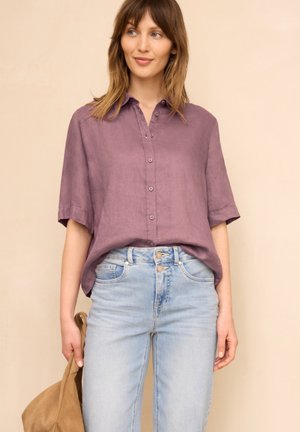 Kobieta z włosami do ramion, ubrana w luźną fioletową koszulę na guziki i jasnoniebieskie dżinsy, trzymająca jasnobrązową kurtkę w lewej ręce.