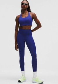 Blå atletisk sportsbh og matchende højtaljede leggings lavet af strækstof. Lysegrønne atletiske sko med hvide accenter.